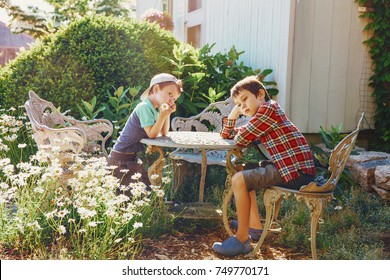 Children Are Sitting In The Garden At The Table. Bored Kids In The Yard Near The House. The Concept Of Idleness And Apathy