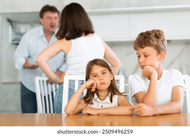 Children are sitting at kitchen table and hear their parents swearing, talking about divorce. Husband and wife are shouting at each other. Behind children are screaming parents in background, blurred - Powered by Shutterstock