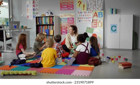 children sitting on floor while caring teacher explains lesson using toy in kindergarten. Elementary school students and teacher sit in circle in classroom