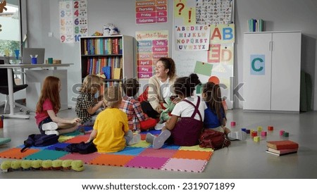 children sitting on floor while caring teacher explains lesson using toy in kindergarten. Elementary school students and teacher sit in circle in classroom