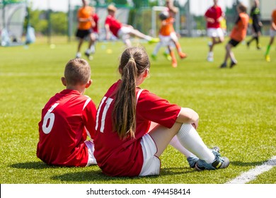 Children Soccer Players Playing Game. Young Girl And Boy Soccer Players Sitting Together On Grass Football Field