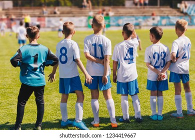 Children Sport Team Photo. Group Of Young Boys Playing Soccer Tournament Match. Football Soccer Game For Children. Kids Soccer Players Standing Back On Bench And Watching Tournament Game