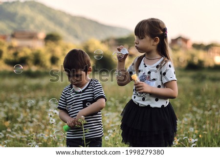 Children are standing side by side in chamomile field, blowing soap bubbles. Charming kids on walk in clearing relax among wild beautiful flowers. Happy childhood in nature.