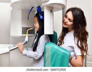 Children Stomatologist Makes Dental Procedure Using  X-ray Machine. Panoramic Radiography For An Asian Child Kid Girl With Special Dentist Equipment In Medical Cabinet