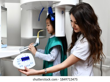 Children Stomatologist Makes Dental Procedure Using  X-ray Machine. Panoramic Radiography For An Asian Child Kid Girl With Special Dentist Equipment In Medical Cabinet