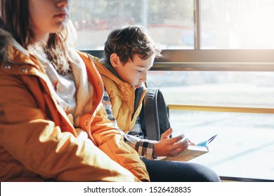 Children Students In Yellow Jackets Riding In The City Bus. Boy Reading A Book At The Window