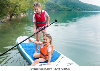 Children In Swim Life Vest Swimming On Stand Up Paddle Board Together. Girls Paddleboarding On SUP Board On The Lake. Active Leisure With Kids. Family Local Getaway Concept