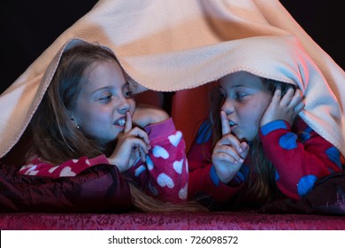 Children And Technology Concept. Kids Wearing Red Jammies In Bed On Black Background. Pyjamas Party For Children. Girls Shushing With Their Fingers On Lips. Girl Friends Play Under Blanket