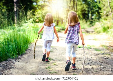 Children - Twin Girls Are Hiking In The Mountains. Active Family And Children Mountaineering In The Nature. Kids Are Walking In Woods With Walking Sticks And Sun In The Background.