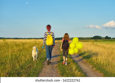 Children Two Girls Walking Along Country Road With White Dog And Yellow Balloons, Back View. Background Sunset, Blue Sky, Summer Meadow