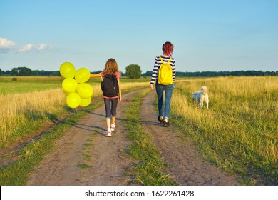 Children Two Girls Walking Along Country Road With White Dog And Yellow Balloons, Back View. Background Sunset, Blue Sky, Summer Meadow