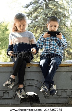 Children use gadgets while sitting in the park outdoors, games on smartphone and tablet.