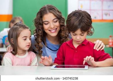 Children Using Digital Tablet With Teacher At Classroom Desk