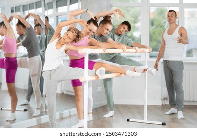 Children work at ballet machine, put their foot on crossbar of bare and stretch leg muscles. Preparatory activities for dance lesson, warm-up - Powered by Shutterstock