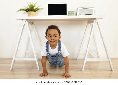 Children, Workout, Fitness And Sports Concept. Adorable Positive African American Schoolboy Dressed In Jeans Jumpsuit Doing Physical Exercises Indoors, Planking On Floor With Feet Under Table, Smiling