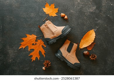 Children's beige boots with black soles on a dark textured background with autumn leaves, acorns, and pinecones. Stylish fall footwear for kids. - Powered by Shutterstock