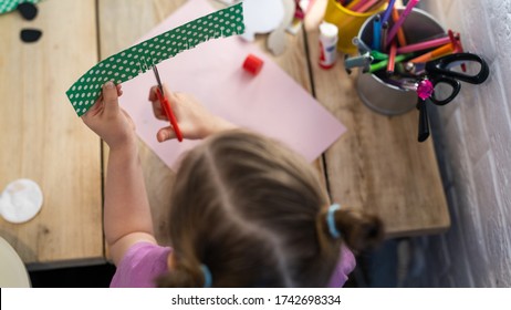 Children's Creativity. A Little Girl Enthusiastically Cuts Colored Cardboard With Scissors, Cutting Small Strips In The Form Of Grass On It. Skill To Cut With Scissors, Learning New Things. Top View