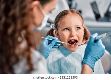 Children's dentistry. First examination at the dentist. A cute beautiful girl with an open mouth is looking to the side while the doctor is treating her teeth. - Powered by Shutterstock
