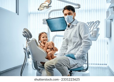 Children's dentistry.Portrait of a dentist doctor in a medical gown and mask sitting and in the background a little female patient sitting in a chair and holding a toy.  - Powered by Shutterstock