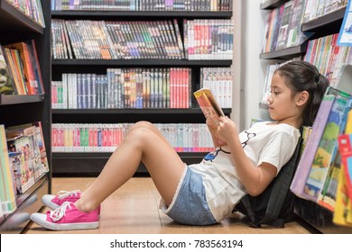 Children's education lifestyle learning concept with school girl kid reading book sitting on the floor of bookstore or library reading cartoon paperback among  bookshelf aisle