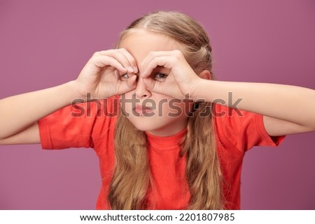 Children's emotions and games. A funny blonde girl with pigtails in a red t-shirt makes a funny face and looks with suspicion through imaginary binoculars. Purple background. 
