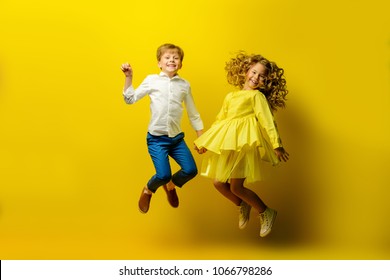 Children's fashion. Beautiful boy and girl in elegant clothes jumping together at studio over yellow background.