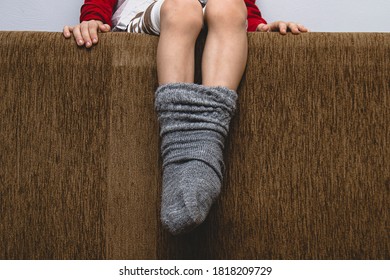 Children's Feet In One Woolen Sock. The Legs Are Frozen, My Mother's Concern Is To Dress The Child. Heating, Cooling, Autumn Concept On Gray Background Copy Space. Mother Tied Son With Her Care