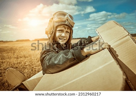Children's games and dreams. A cute kid in a leather jacket and aviator's helmet plays in a wheat field with a cardboard plane and looks curiously into the distance. Sunny day.