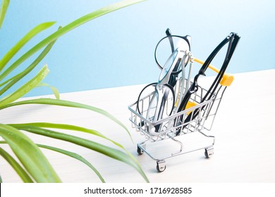 Children's Glasses And Two Pairs Of Black Glasses For Adults Are In A Small Shopping Trolley On A Blue Background, Copy Space, Online Purchase Of Glasses, A Flower In The Foreground In Blur