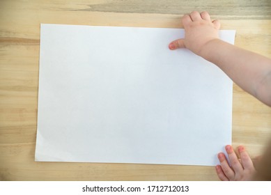 Childrens Hand Holding Blank White Paper Sheet On Wooden Background. Copy Space. Template. Mockup.