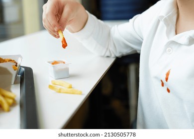 Children's Hand Holding  French Fries Potato Chips And Tomato Sauce At The Table In A Fast Food Restaurant. Dirty Ketchup Stains On White Kid Clothes. 