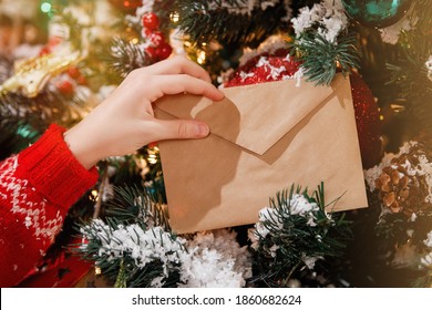 Children's hand holds an envelope with a letter to Santa on Christmas Eve against the background of a decorated Christmas tree with bright lights. Selective focus.