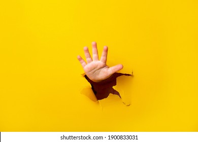 Children's Hand Sticks Out Of A Hole In A Sheet Of Paper On A Yellow Background. Five Finger Sign, Copy Space.