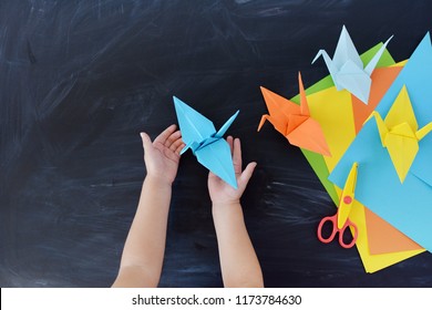 Children's Hands Do Origami From Colored Paper. Lesson Of Origami. Color Paper Cranes On A Black Background. Creativity. The Kid's Hands 