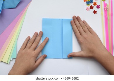 Children's Hands Do Origami On A Table. Nearby Sheets Of Color Paper Lie.