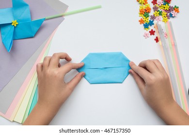 Children's Hands Do Origami On A Table. Nearby Sheets Of Color Paper Lie.