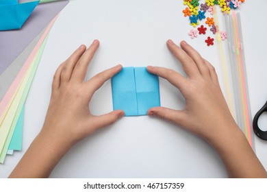 Children's Hands Do Origami On A Table. Nearby Sheets Of Color Paper Lie.