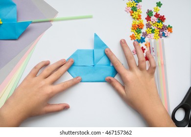 Children's Hands Do Origami On A Table. Nearby Sheets Of Color Paper Lie.