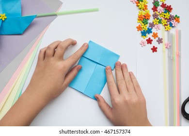 Children's Hands Do Origami On A Table. Nearby Sheets Of Color Paper Lie.