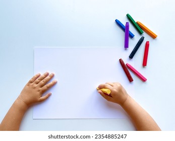 Childrens hands draw with colored wax crayons on a white sheet of paper. Top