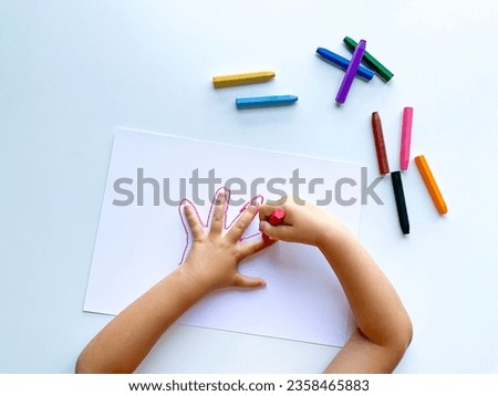 Childrens hands draw their hand with wax crayons on white paper, top view.