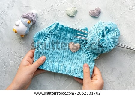 Children's hands holding a knitted T-shirt for a toy made of turquoise cotton yarn. The development of fine motor skills. Knitting school. Flat lay. A ball of yarn and knitting needles. Hobbies.