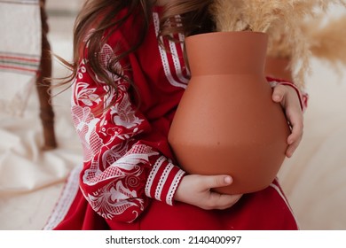 Children's hands hug a clay jug on the background of a traditional embroidered dress