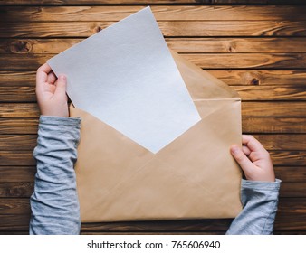 Children's Hands Open A Large Brown Envelope And Take A Clean White Sheet Of Paper Out Of It. Wooden Background.