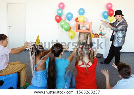 At a children's party, a magician, an illusionist shows tricks to a group of children in caps and with balloons.