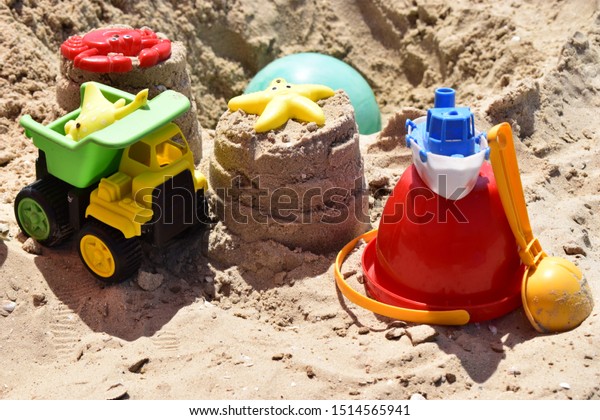 Children\'s plastic toys green end yellow car,\
shovel, red bucket, green ball with yellow sand on the beach by\
sea. Children\'s beach toys on sand on a sunny day. Sandbox on the\
playground for games