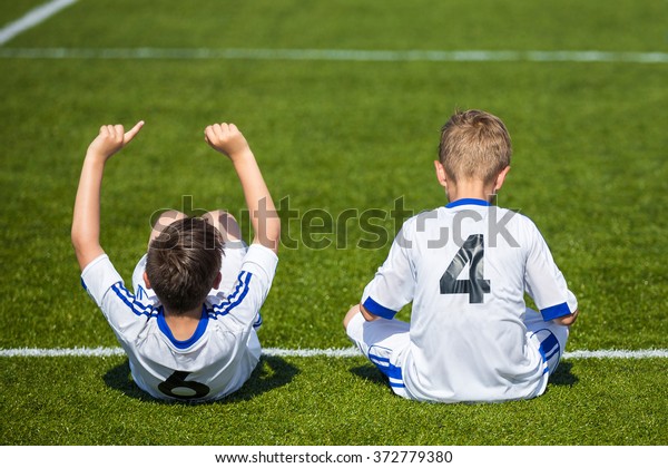 Children's soccer
match. Young boys reserve soccer players sitting on a sport field
and watching football match ready to play. White uniforms of soccer
players with numbers on back.
