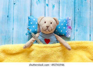 children's soft toy teddy bear in bed with thermometer and pills, take the temperature of a mercury glass . On  blue wooden background   yellow blanket. Playing in hospital.