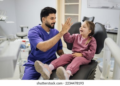 Children's teeth treatment. Smiling little girl sitting in the dentist's chair in light modern dental clinic and excited confident man dentist giving high five. - Powered by Shutterstock