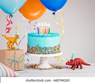 Childs birthday party scene with blue cake, gift box, toy dinosaurs, hats and colorful balloons over light grey.